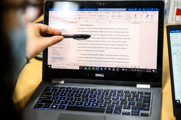 A laptop with a paper displayed and a hand going through the essay line by line